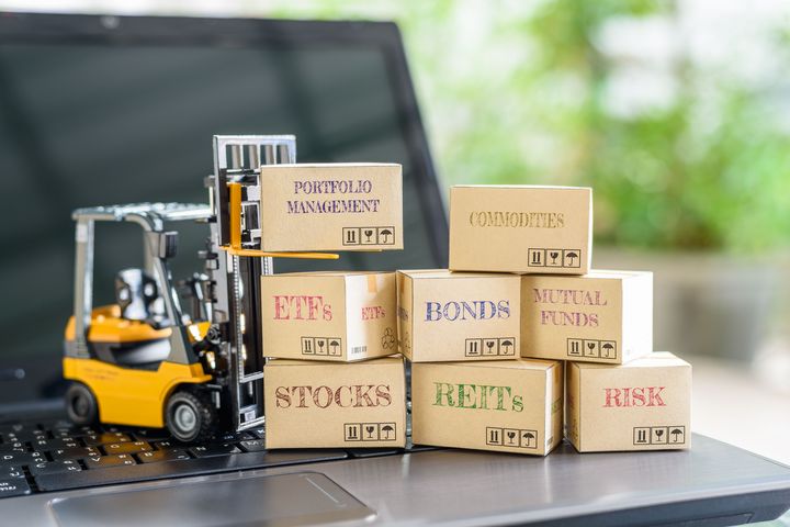 Risiken über verschiedene Anlageklassen streuen Miniatur-Gabelstapler vor einem Laptop stapelt Kartons mit den Aufschriften Aktien, Anleihen, ETFs, Rohstoffe, Fonds, Immobilienanlagen und Risiko.