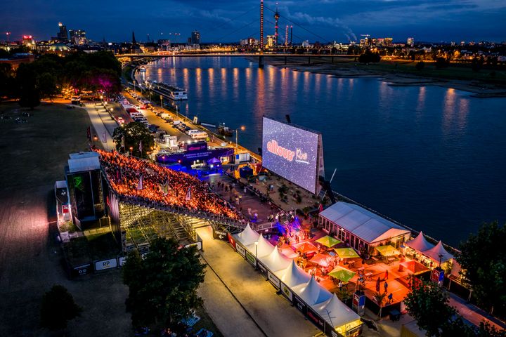alltours Kino im Rheinpark Düsseldorf Blick von oben auf das alltours Kino im Rheinpark Düsseldorf bei Nacht mit großer Leinwand, Publikum, Rhein, Oberkasseler Brücke und beleuchteter Skyline.