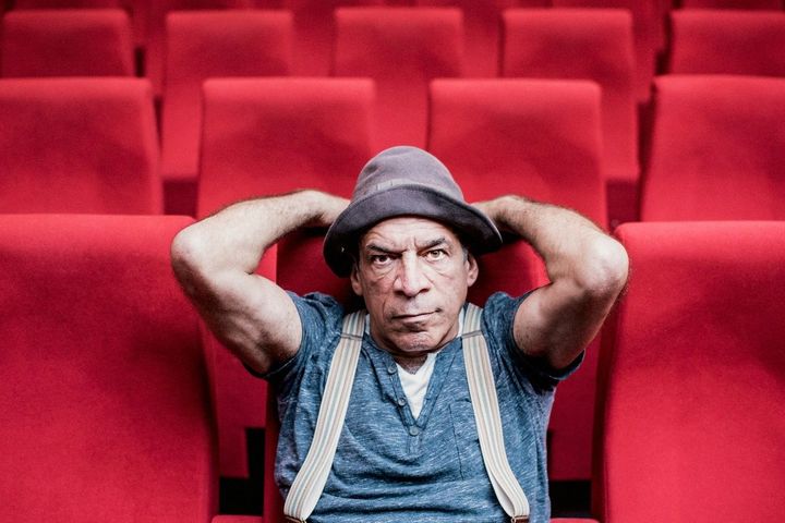Peter Shub im roten Theatersaal Peter Shub sitzt in einem roten Theatersaal, trägt Hut und Hosenträger und blickt frontal in die Kamera.